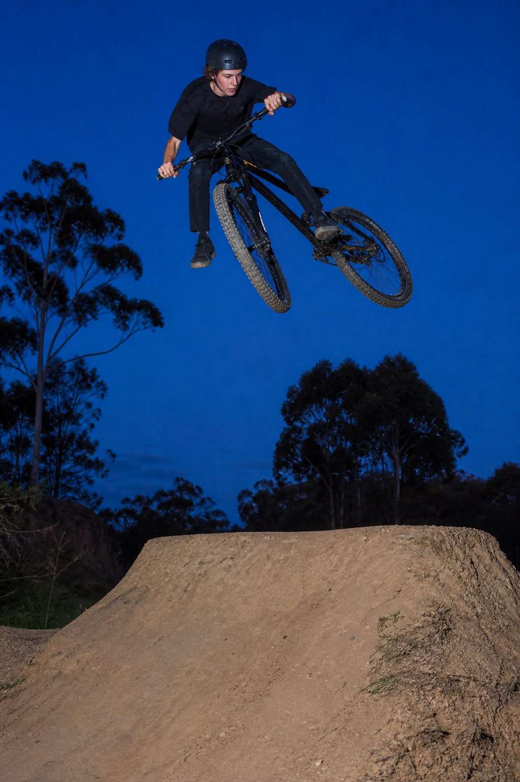 A rider clearing a dirt jump against a deep blue evening sky